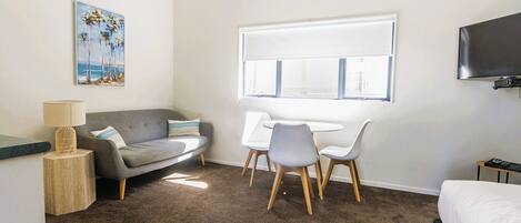A hotel room with a grey sofa, a small table with a lamp, a desk with two chairs, a bed, and a TV mounted on the wall.