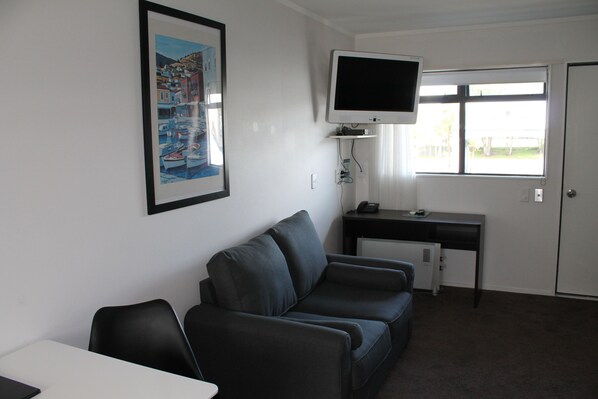 A room with a black leather sofa, a desk with a computer, a flat-screen TV, and a window with a view of a residential area. - Aquarius Motor Inn (Tauranga)