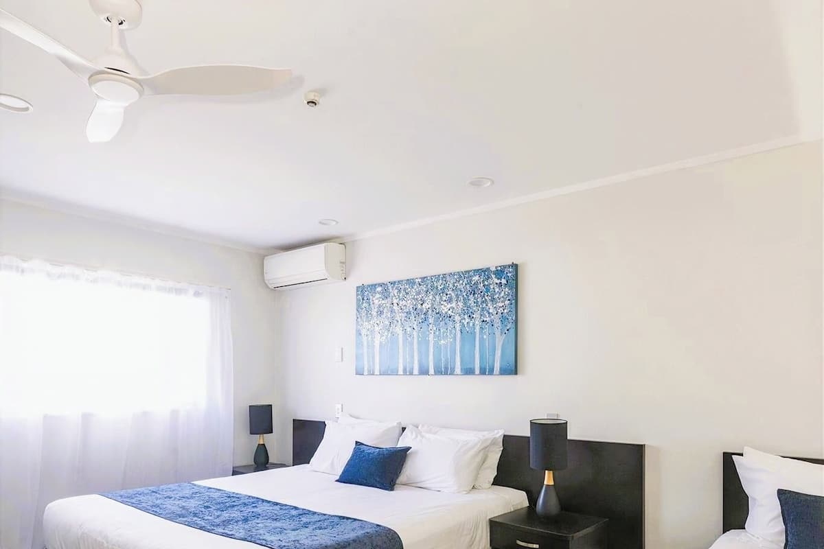 A hotel room with two beds, a ceiling fan, an air conditioning unit, a painting, and a nightstand with a lamp.