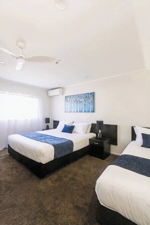A hotel room with two beds, a ceiling fan, an air conditioning unit, a painting, and a nightstand with a lamp. - Aquarius Motor Inn (Tauranga)