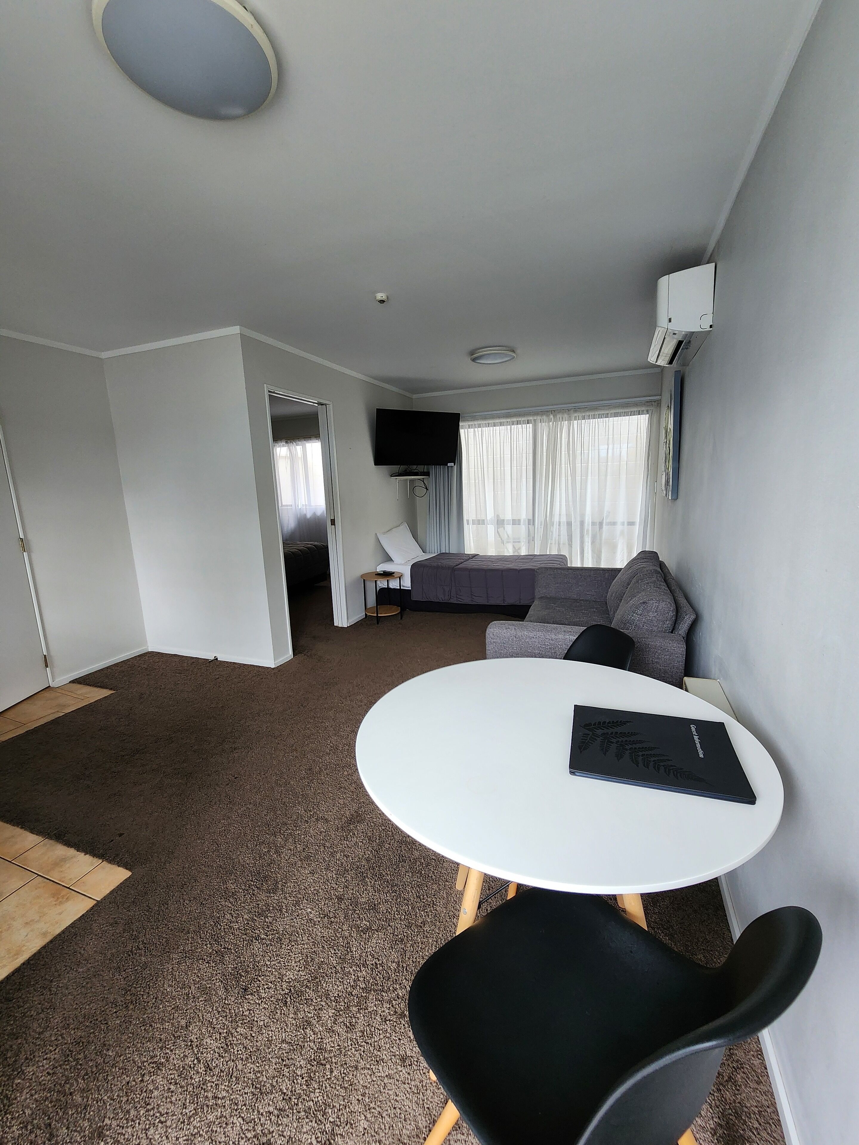 Una habitación moderna y luminosa con techo blanco, una mesa redonda blanca con una silla negra, un sofá gris y un televisor fijado a la pared.