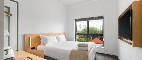 A hotel room with a bed, a desk with a lamp, a television, and a window with a view of buildings and trees.