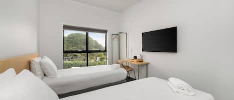 A hotel room with two beds, a desk, a chair, a flat-screen TV, and a window with a view of greenery.