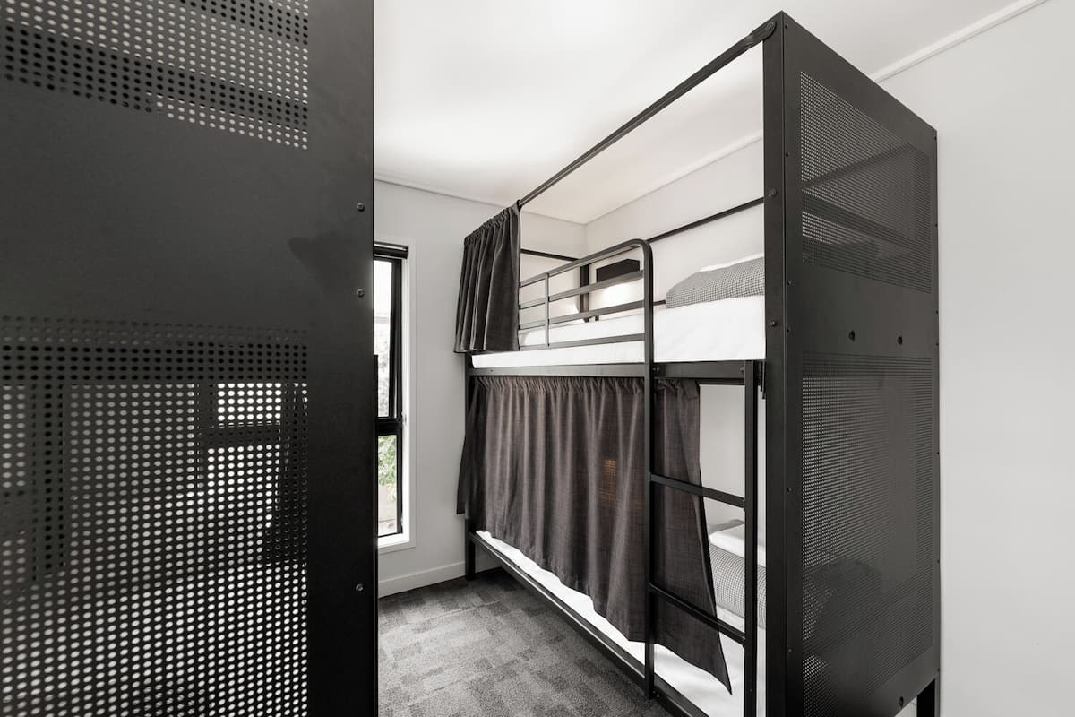 Une chambre avec un lit superposé doté d’une structure en métal, une fenêtre avec des rideaux et une porte munie d’une grille métallique.
