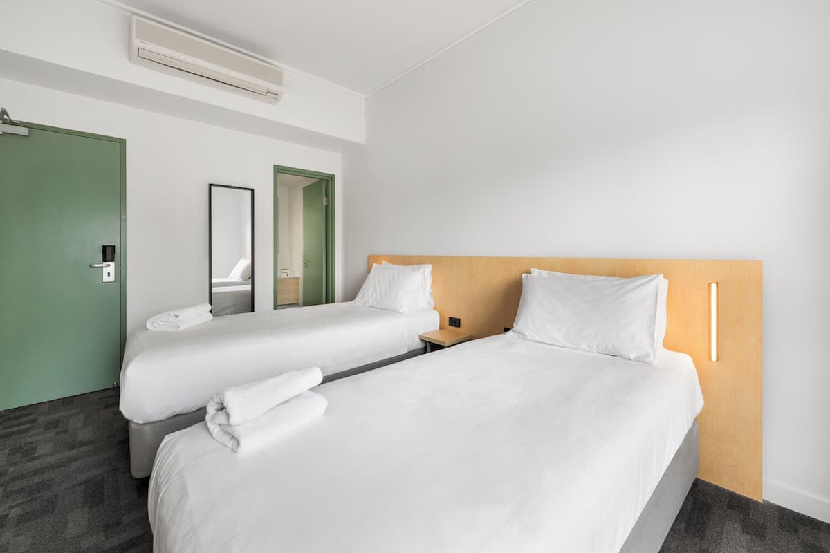 Une chambre d’hôtel avec deux lits simples, une porte verte, un miroir et un climatiseur fixé au mur.