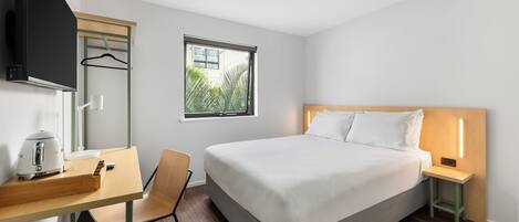 Una habitación de hotel moderna con una cama, mesita de noche, silla, escritorio con hervidor, televisión y ventana con vistas a zonas verdes.
