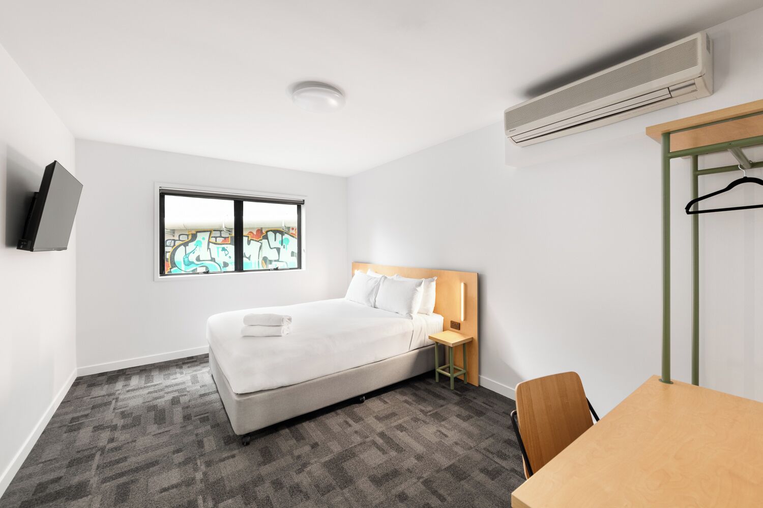 Una habitación de hotel con cama, mesita de noche, silla, televisor, ventana con vistas y unidad de aire acondicionado.