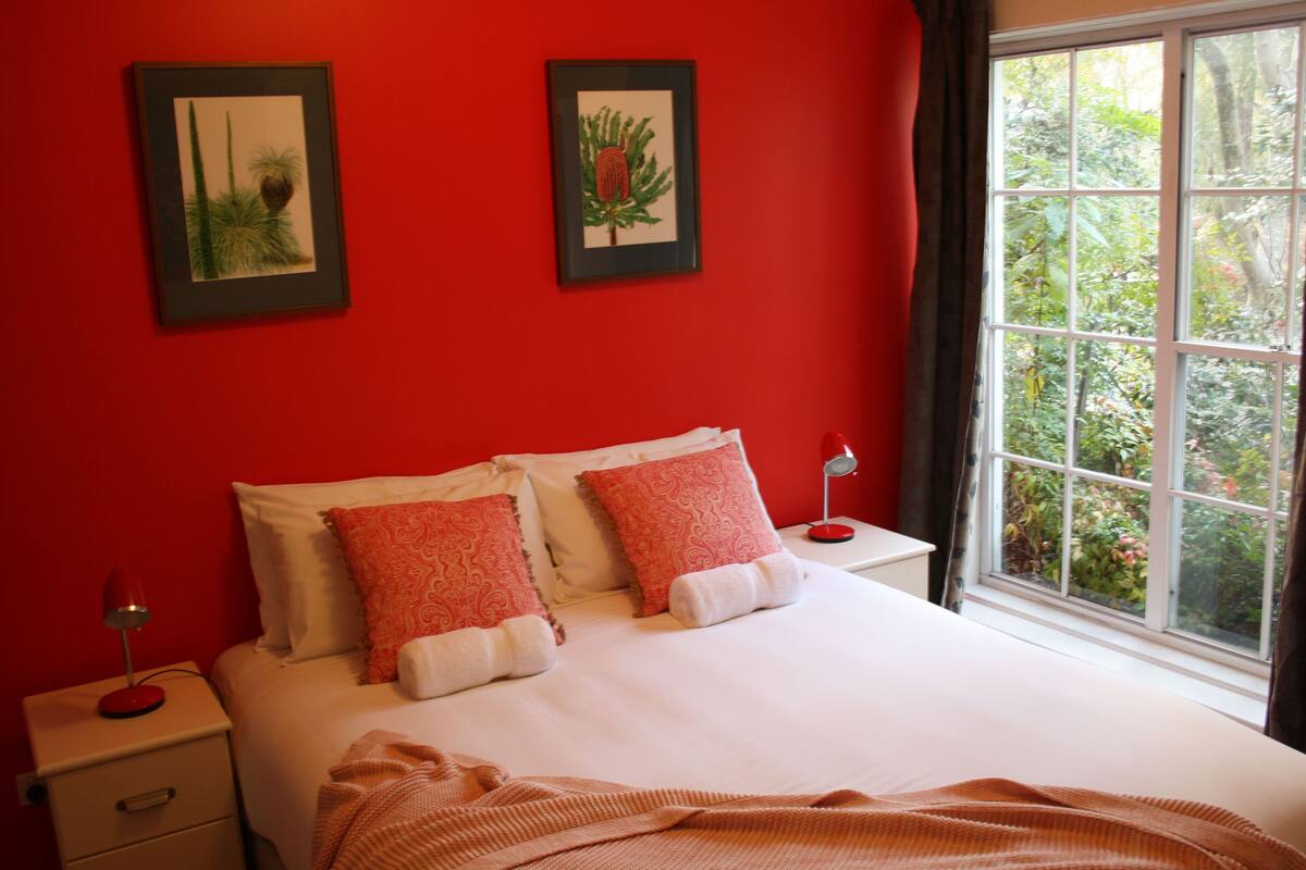 Un dormitorio con pared roja, dos cuadros enmarcados, una cama con ropa blanca y almohadas rojas, una mesita de noche con una lámpara y una ventana con vistas a follaje.