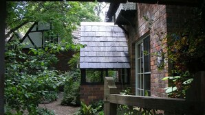 A rustic brick building with a sloped roof, a wooden fence, and lush greenery. - Merrimeet Cottages (Bright)