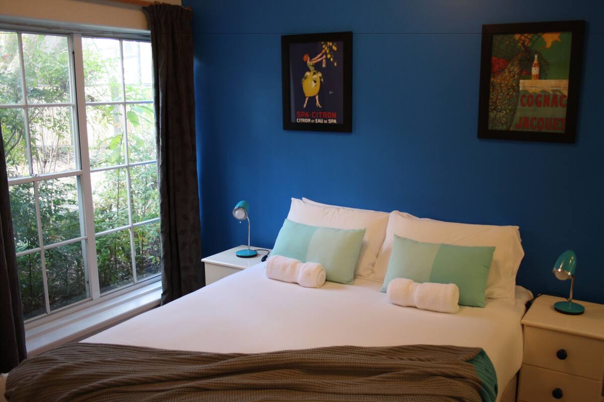 Un dormitorio con una cama grande, dos mesitas de noche, un ventanal con cortinas y dos cuadros colgados en la pared.