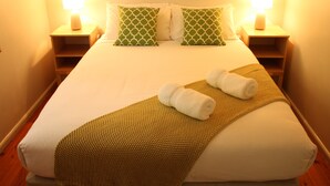 A neatly made bed with a green patterned pillow, a brown textured blanket, and two rolled towels. - Merrimeet Cottages (Bright)