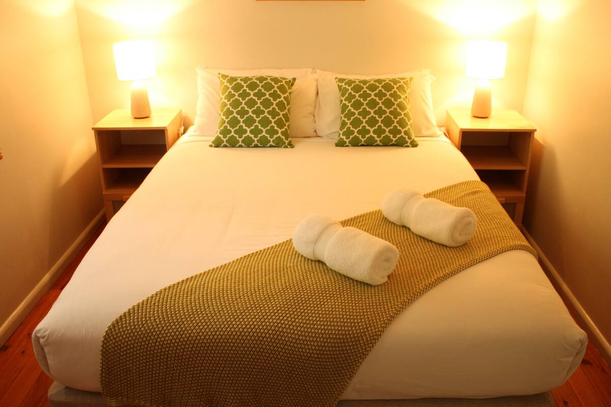 Una cama bien hecha con un cojín estampado en verde, una manta texturizada marrón y dos toallas enrolladas.