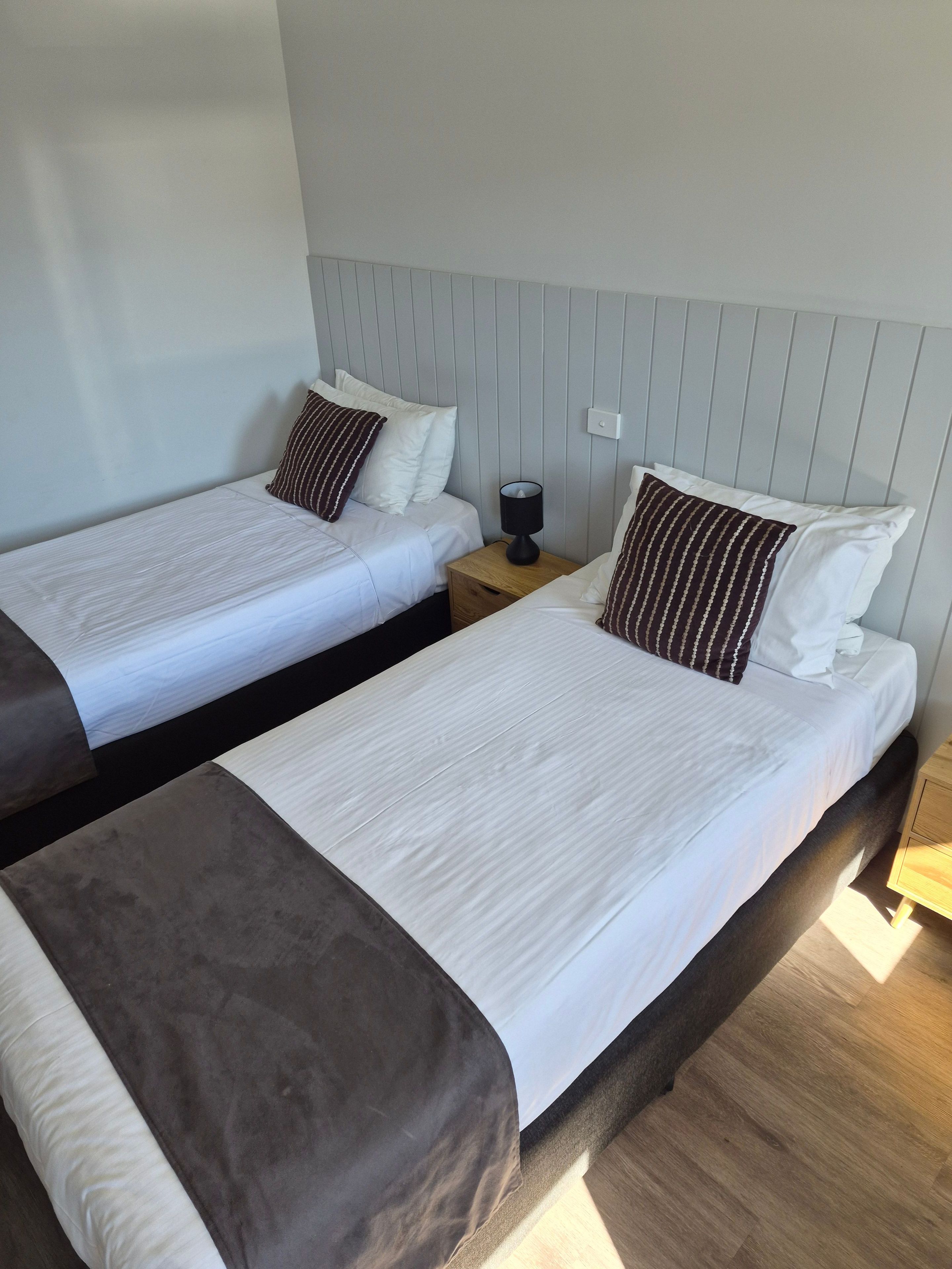 Two single beds with white linens and dark brown headboards, a bedside table with a lamp, and a light-colored wall.