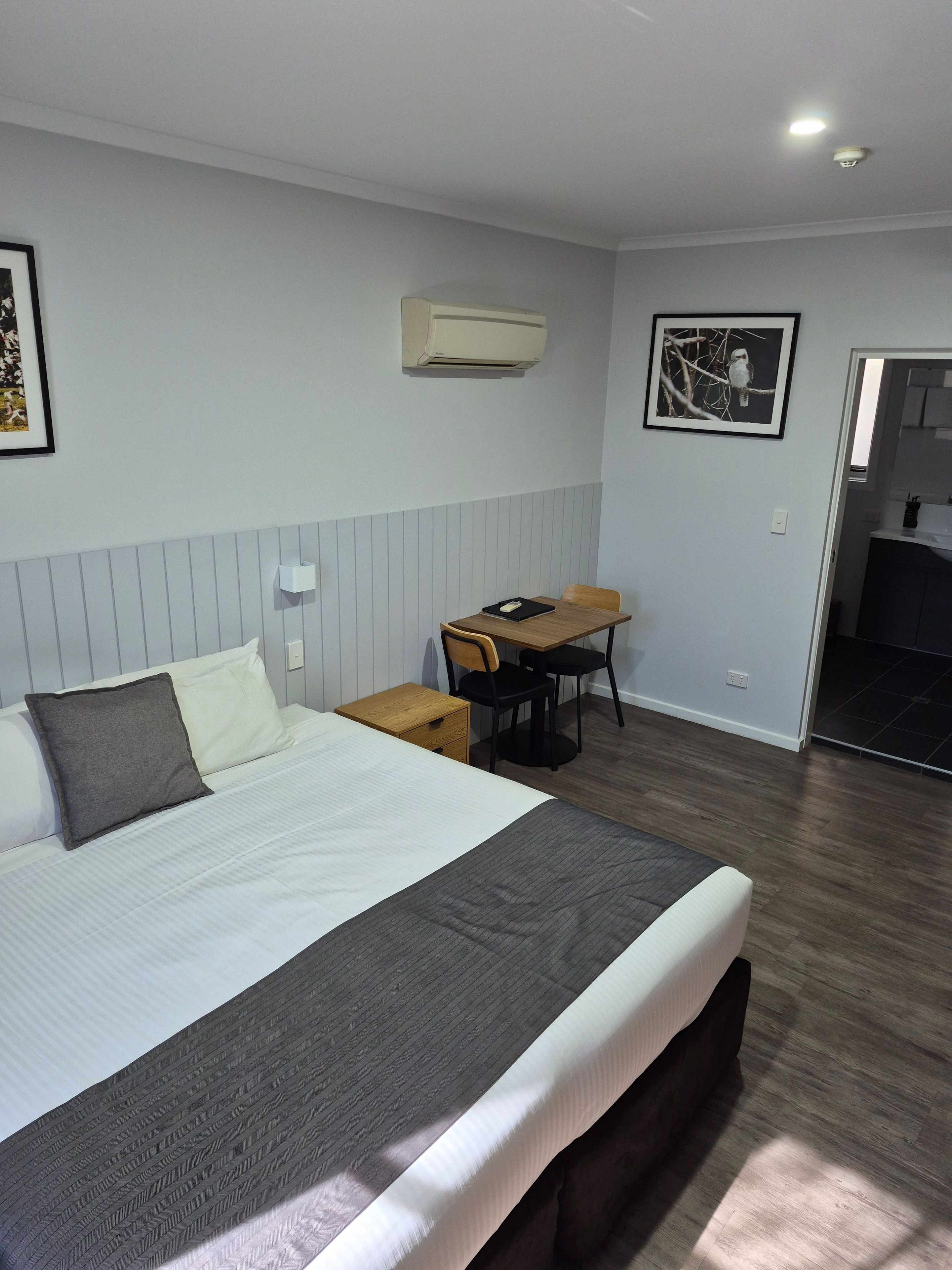 A hotel room with a bed, a desk, a chair, a picture on the wall, and an air conditioning unit.