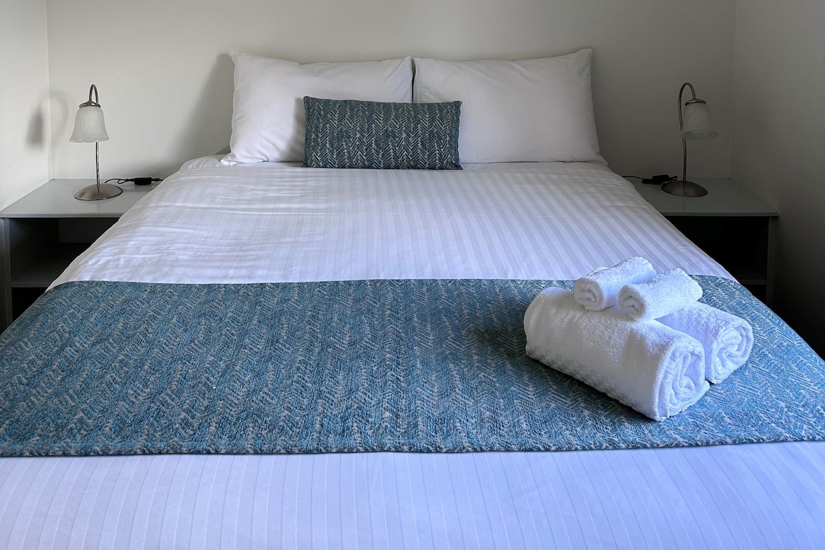 A hotel room with a bed, two bedside tables, a painting of a beach, and a blue patterned bedspread.