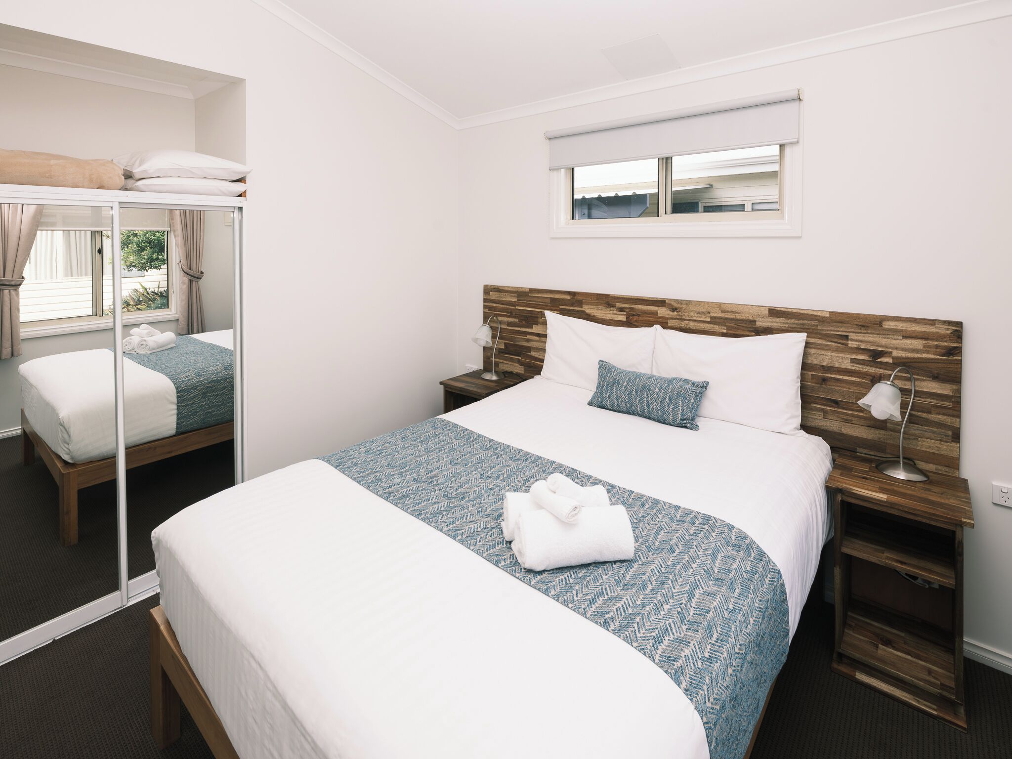 A hotel room with a large bed, a bedside table with a lamp, a window with blinds, and a mirror.