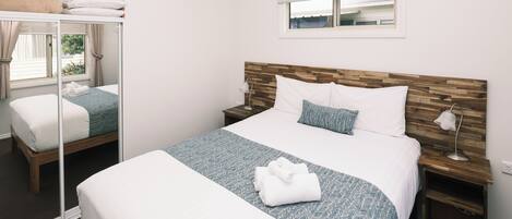 A hotel room with a large bed, a bedside table with a lamp, a window with blinds, and a mirror.