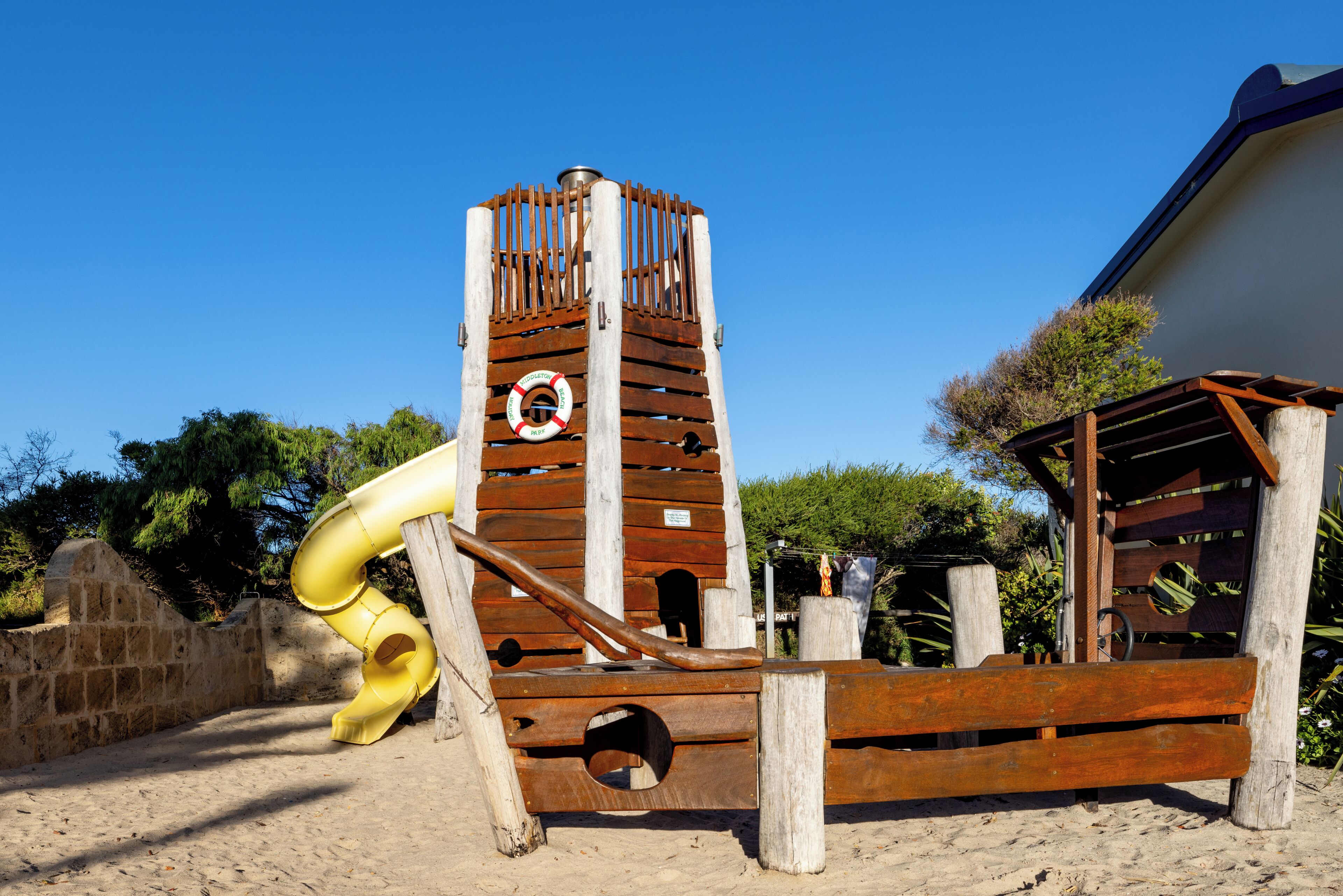 A wooden play structure with a slide and a tower, surrounded by sand and greenery.