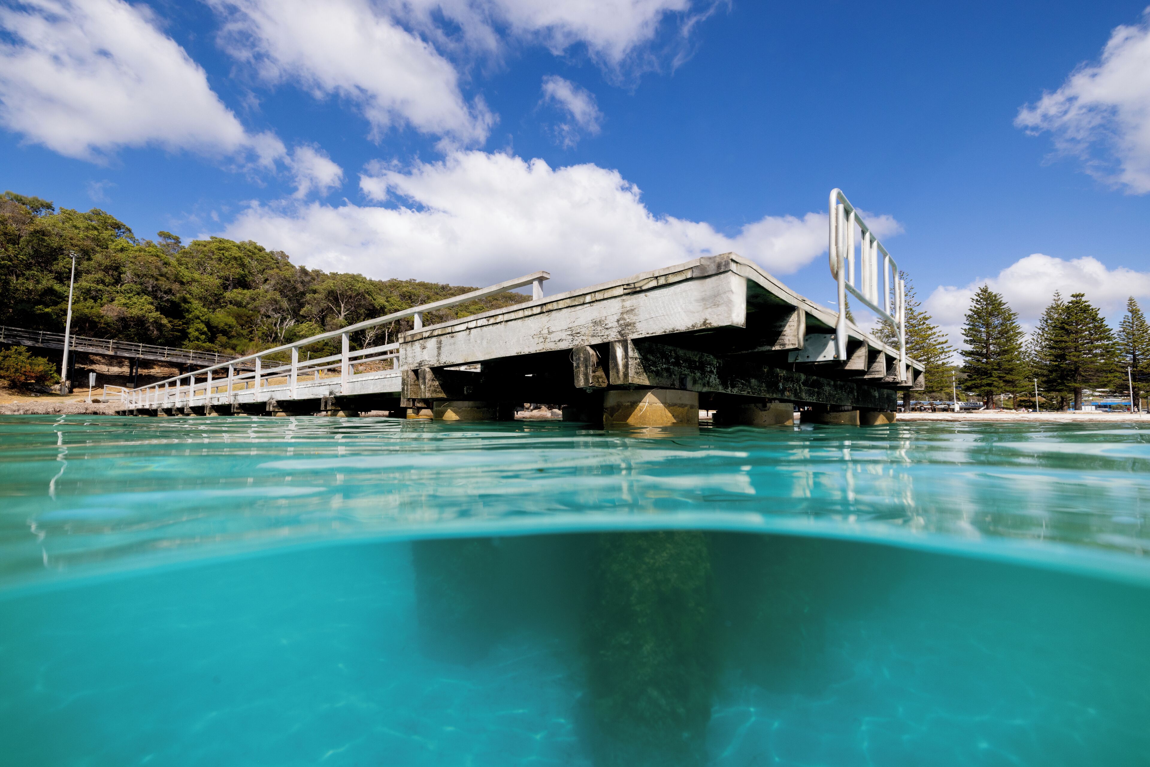 A bridge over a turquoise body of water with a clear sky and trees in the background.