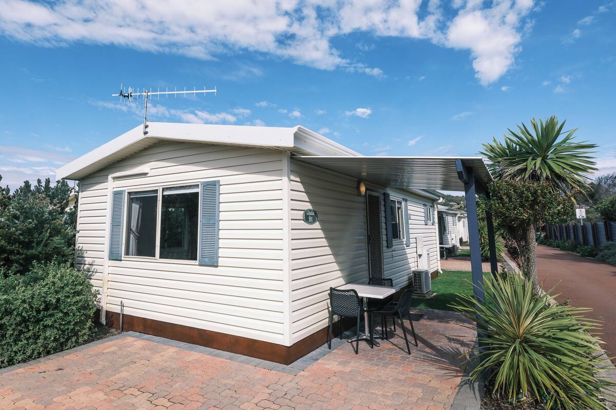 A single-story mobile home with a patio area, a table with chairs, and a small garden with a palm tree.