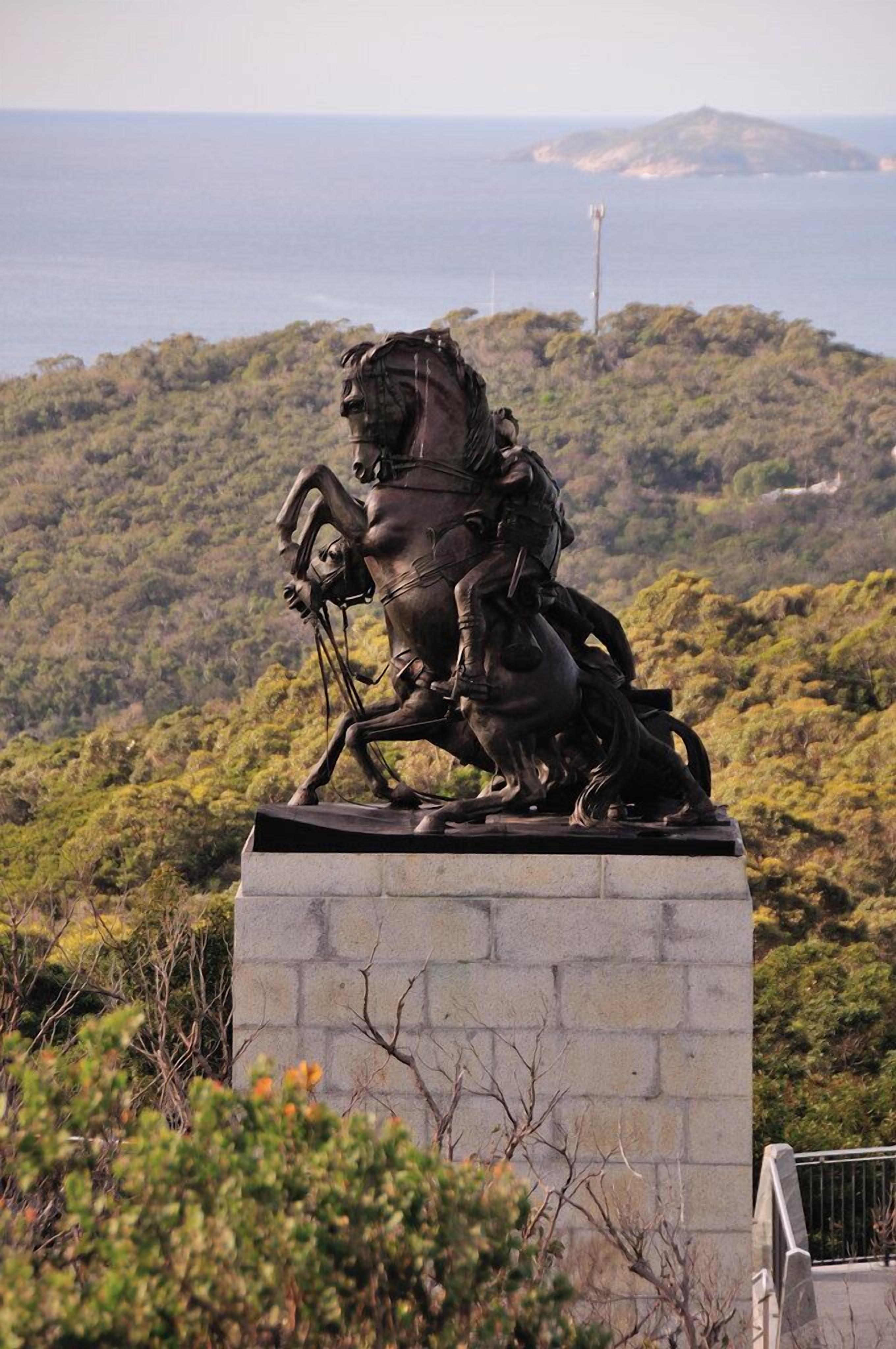A statue of a figure on horseback, set on a stone pedestal, overlooking a coastal landscape with hills and a lighthouse in the distance.