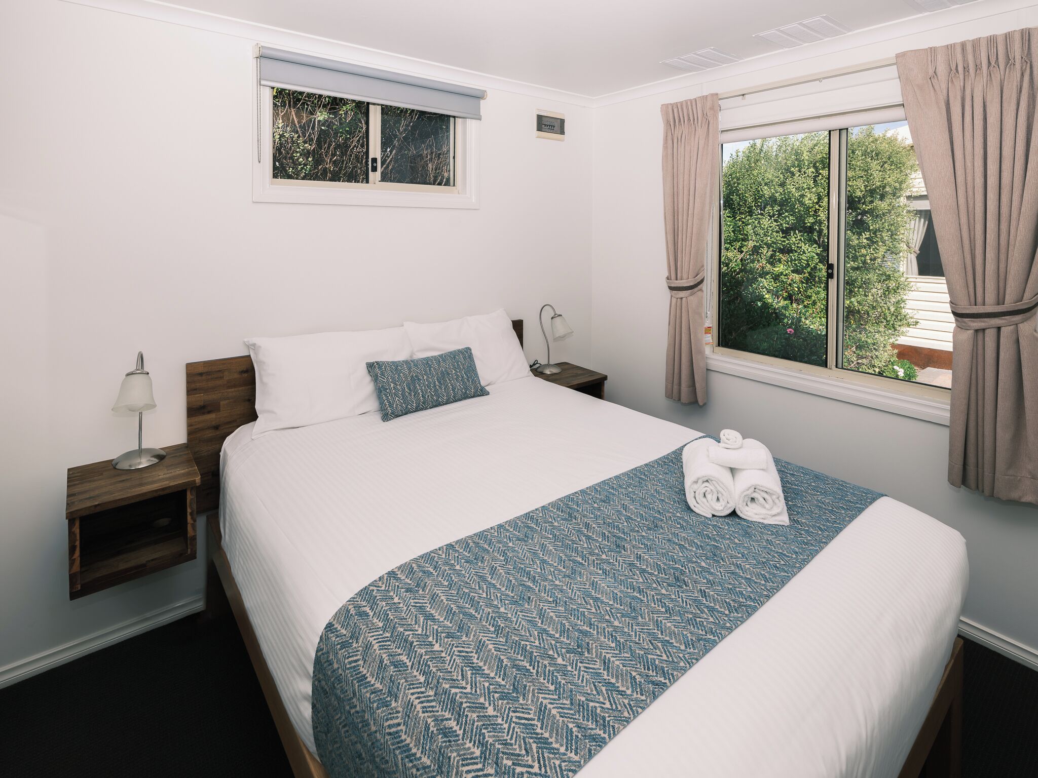 Una cama bien hecha con una colcha azul estampada, un cabecero de madera, una mesita de noche con una lámpara, una ventana con cortinas y vistas al exterior con vegetación.