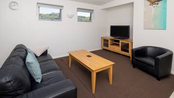 Una sala de estar con un sofá de cuero negro, una mesa de centro de madera, un sillón negro, un televisor sobre un mueble de madera y dos ventanas con vistas a follaje.