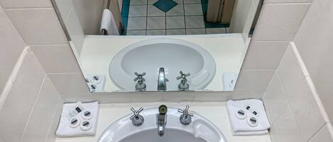 Un baño con lavamanos blanco, dos grifos, un espejo y un dispensador de papel higiénico.