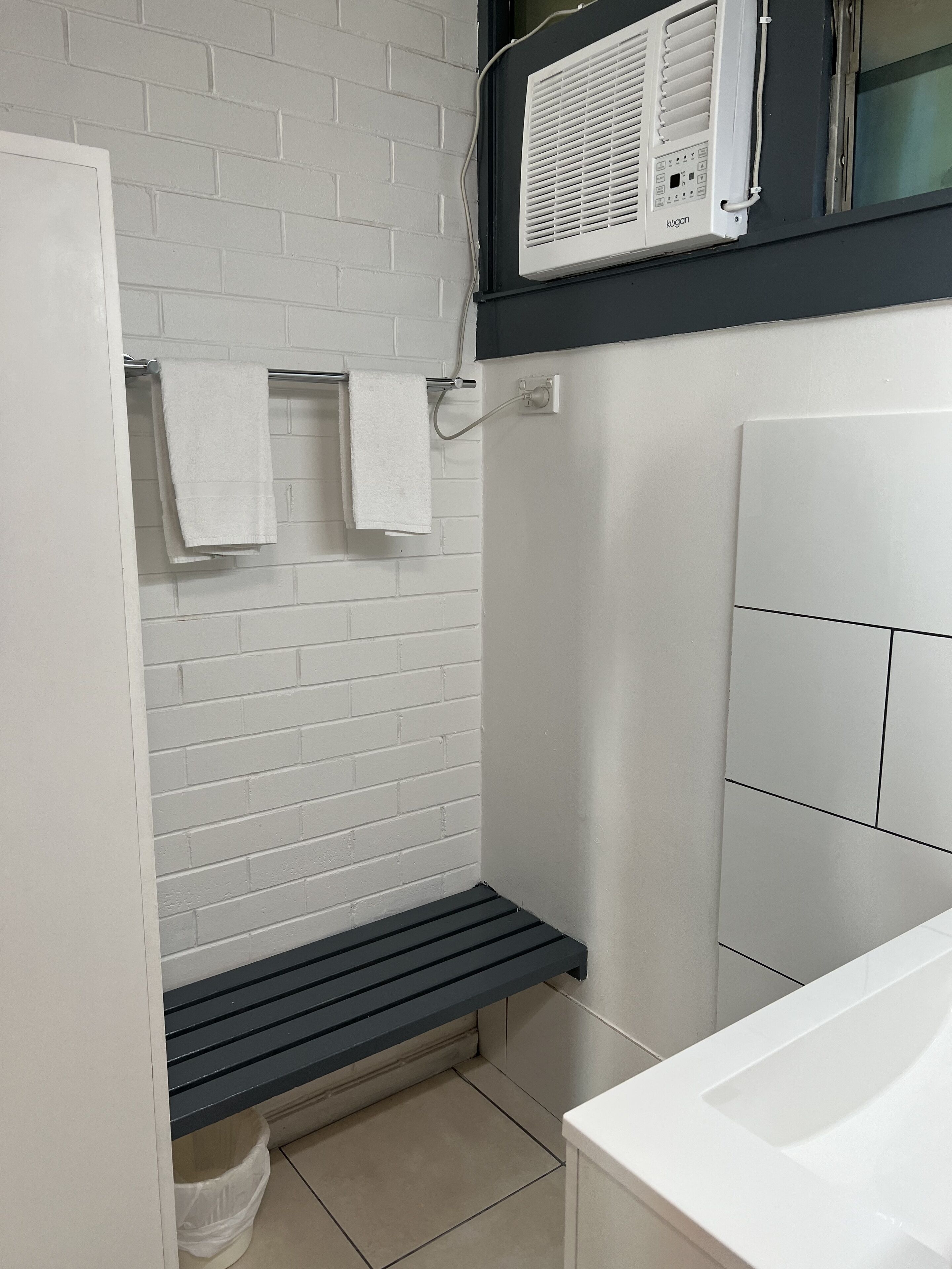 Un baño con pared de ladrillo blanco, un perchero con dos toallas, un banco negro, un lavamanos blanco y una ventana con persianas.