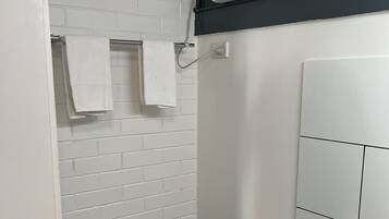 Un baño con pared de ladrillo blanco, un perchero con dos toallas, un banco negro, un lavamanos blanco y una ventana con persianas.