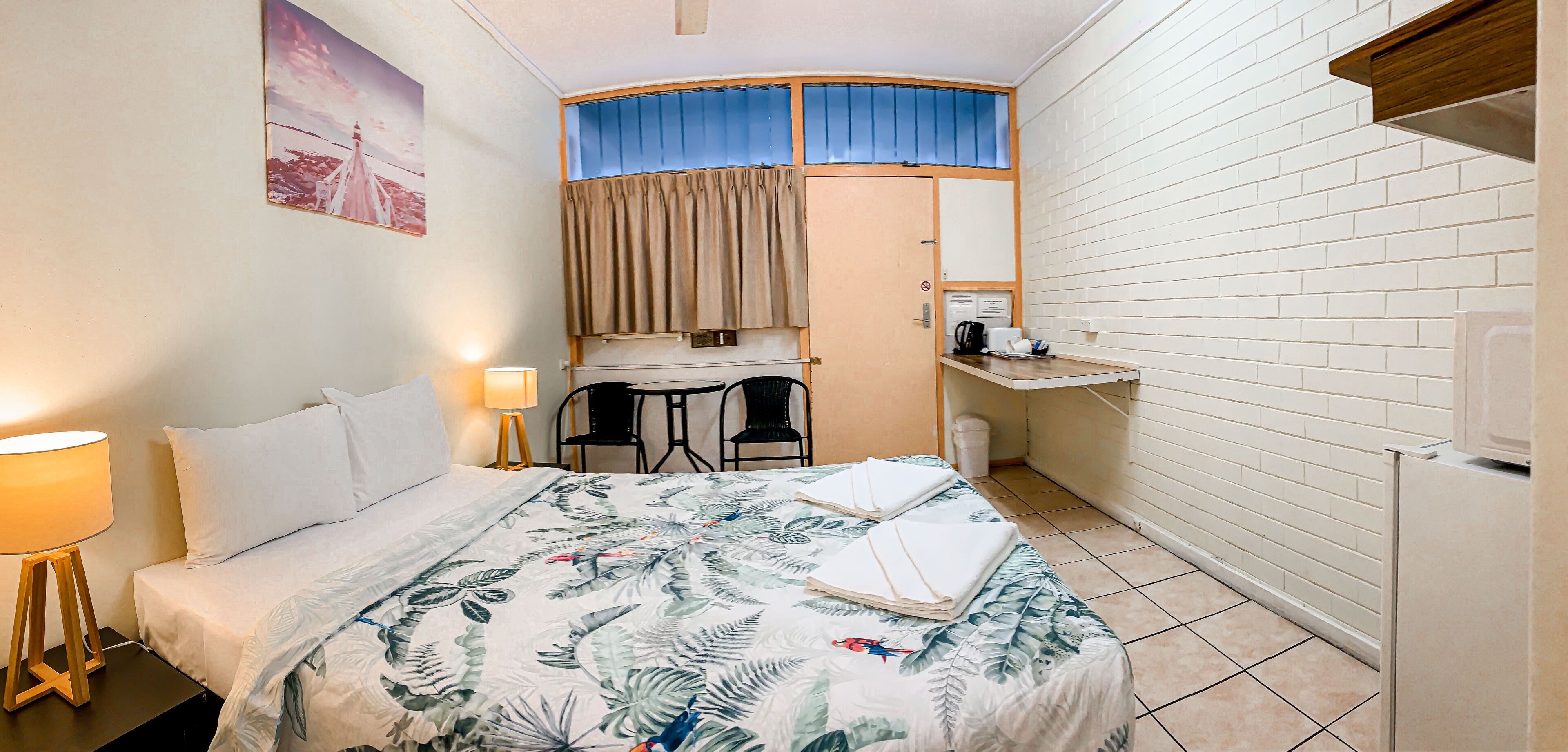 Et lille hotelværelse med seng, skrivebord, to stole, et lille bord, en lampe og et køleskab.