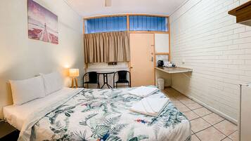 Et lille hotelværelse med seng, skrivebord, to stole, et lille bord, en lampe og et køleskab.