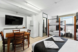 Habitación de hotel compacta con cama, escritorio con sillas, televisor, refrigerador y literas.