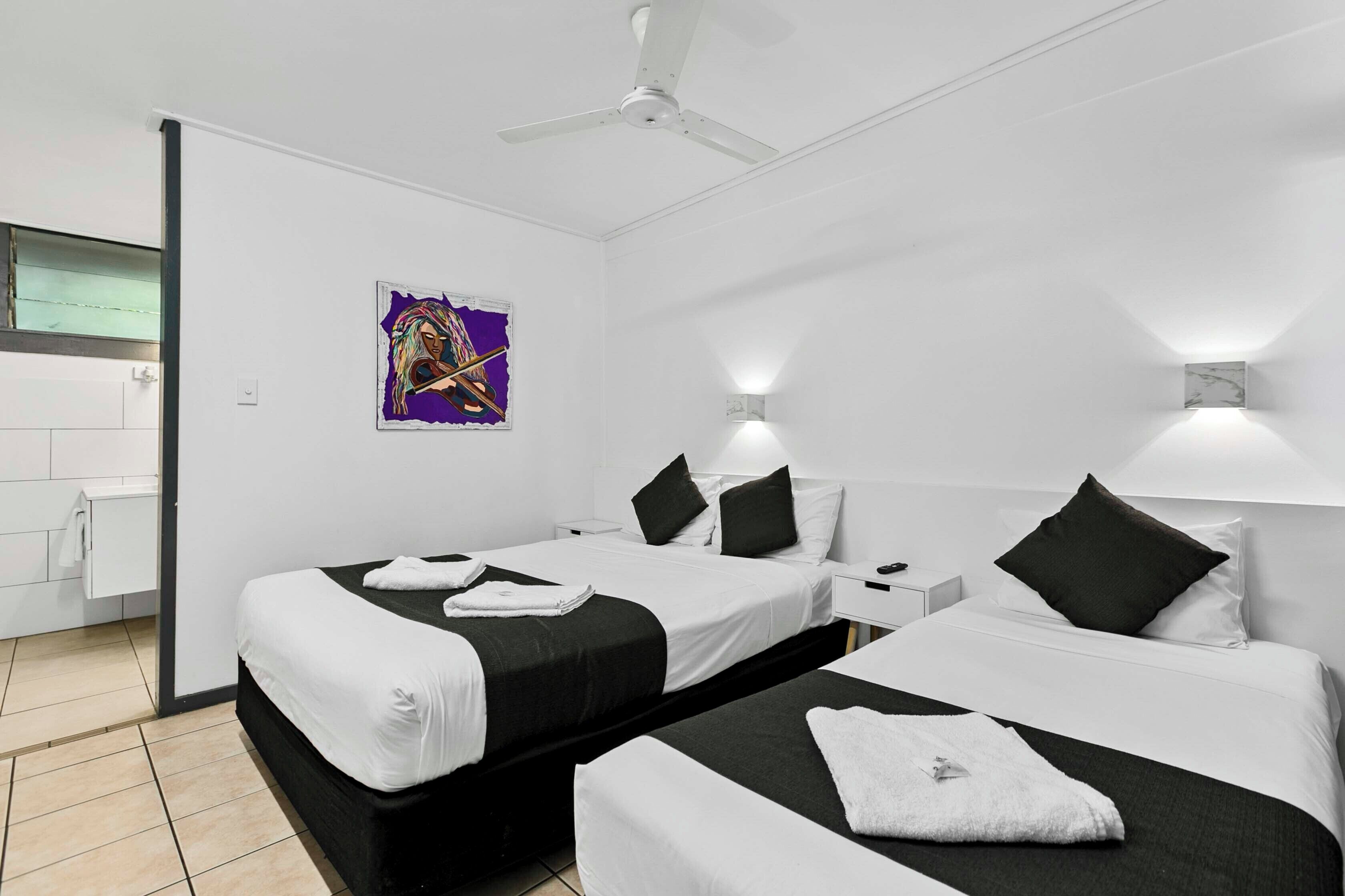 A hotel room with two beds, a ceiling fan, a bathroom with a mirror, and a poster on the wall.