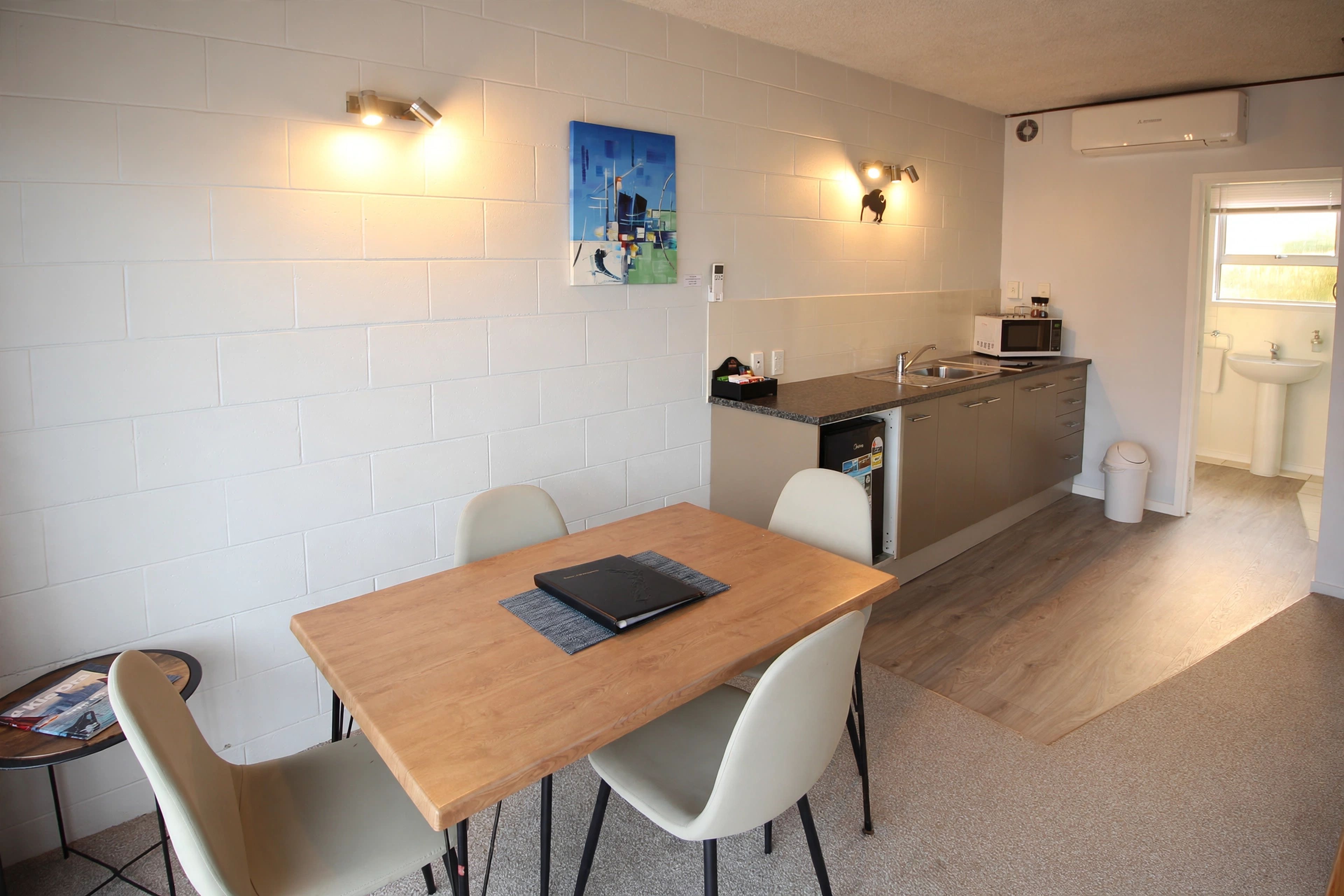 A compact kitchen with a microwave, sink, and a small island. There is a dining area with a table and chairs, and a painting on the wall.