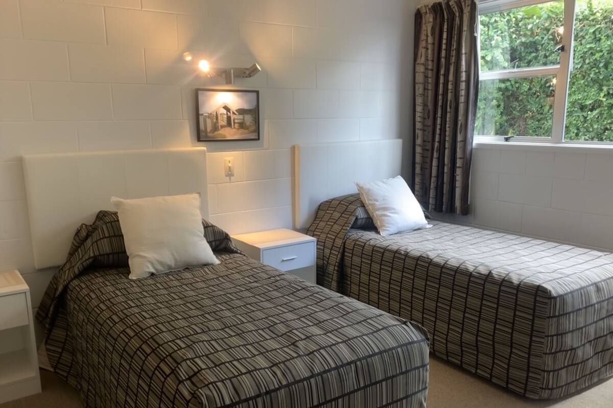 A double room with two beds, each with a striped bedspread, a nightstand, a bedside table, a wall-mounted light, and a window with curtains.