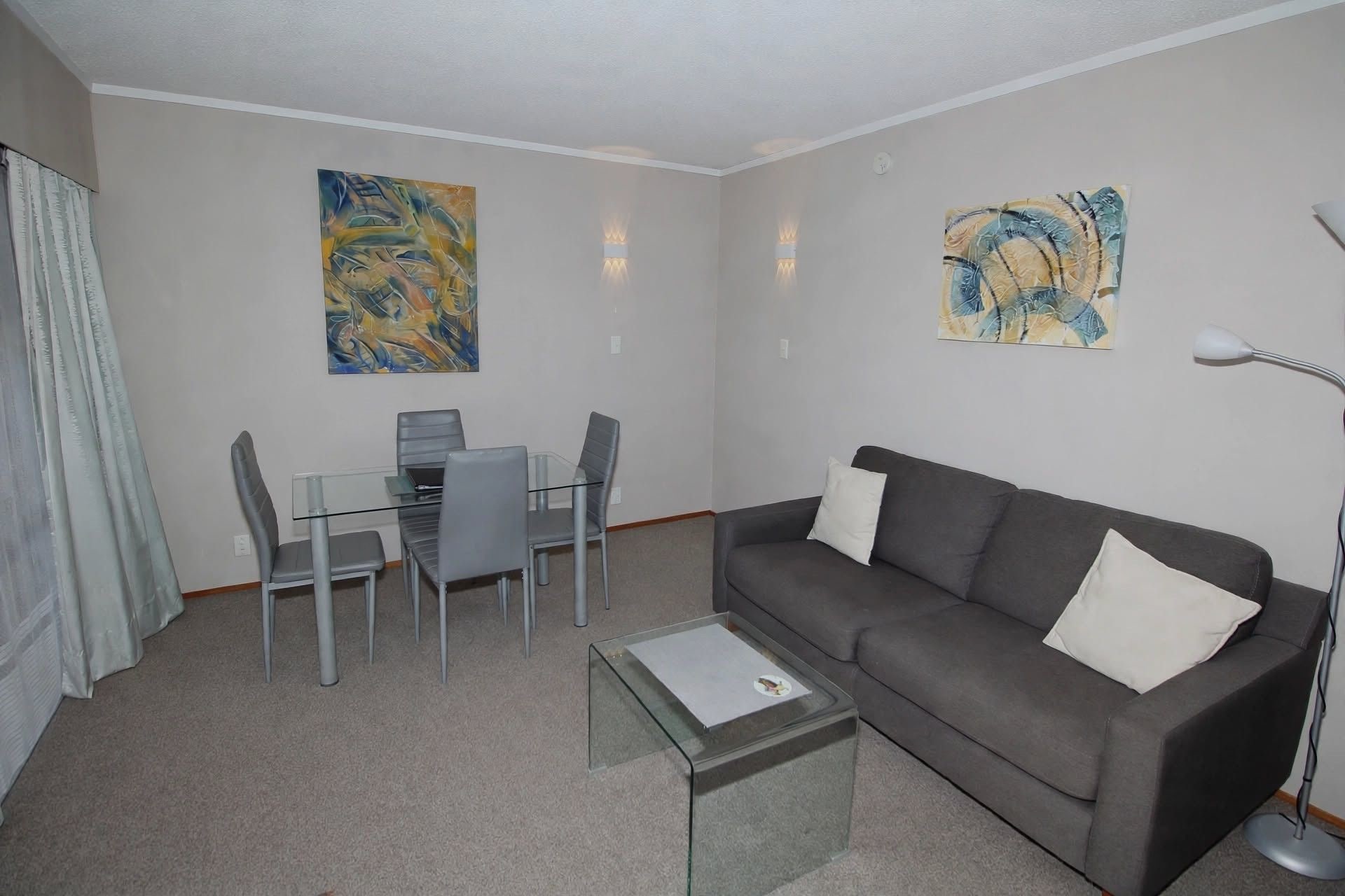 Una sala de estar con un sofá gris, una mesa de centro de vidrio y un comedor con una mesa y sillas de vidrio.