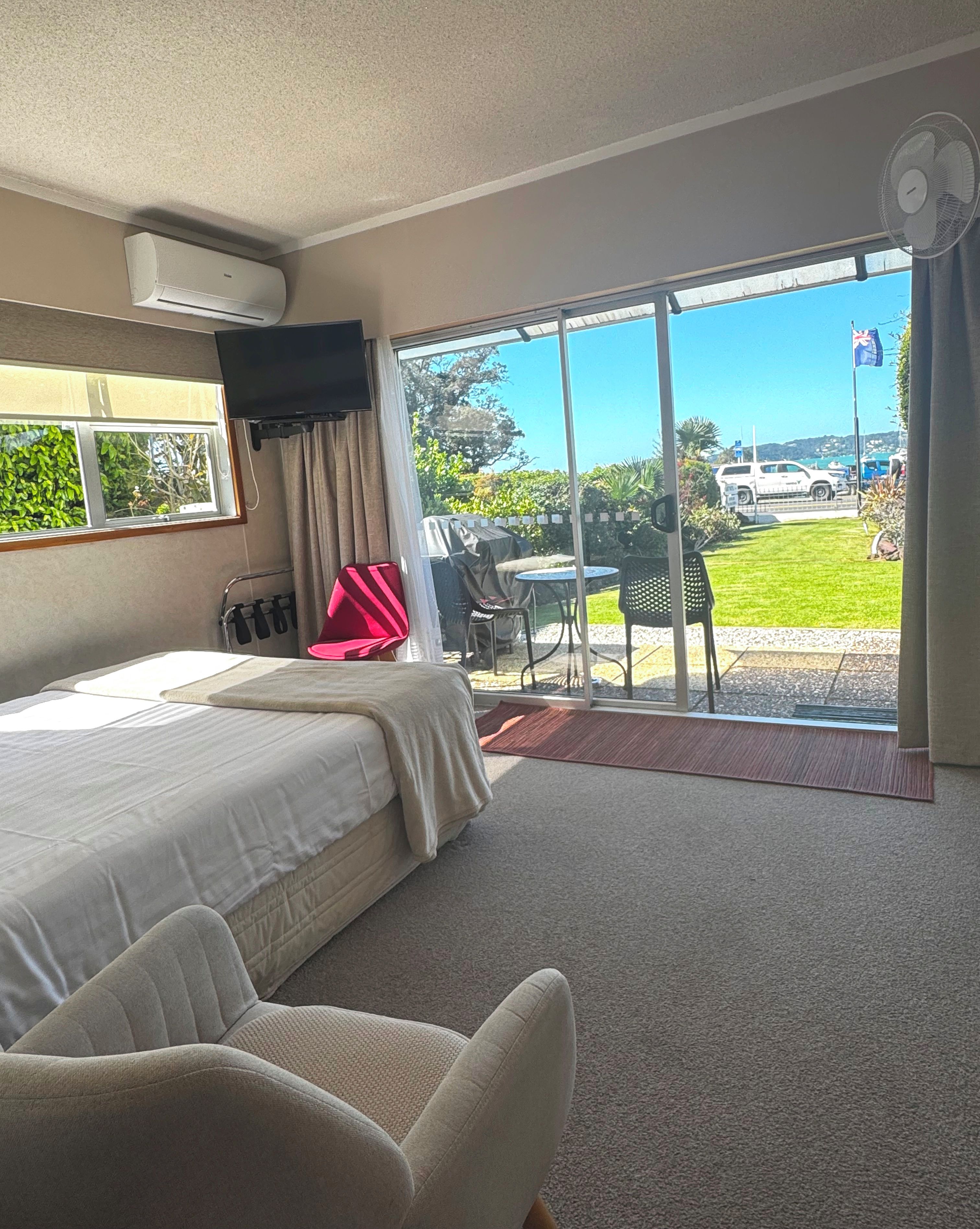 A hotel room with a bed, a TV, a sliding glass door to a patio with outdoor furniture, and a view of the sea and boats.