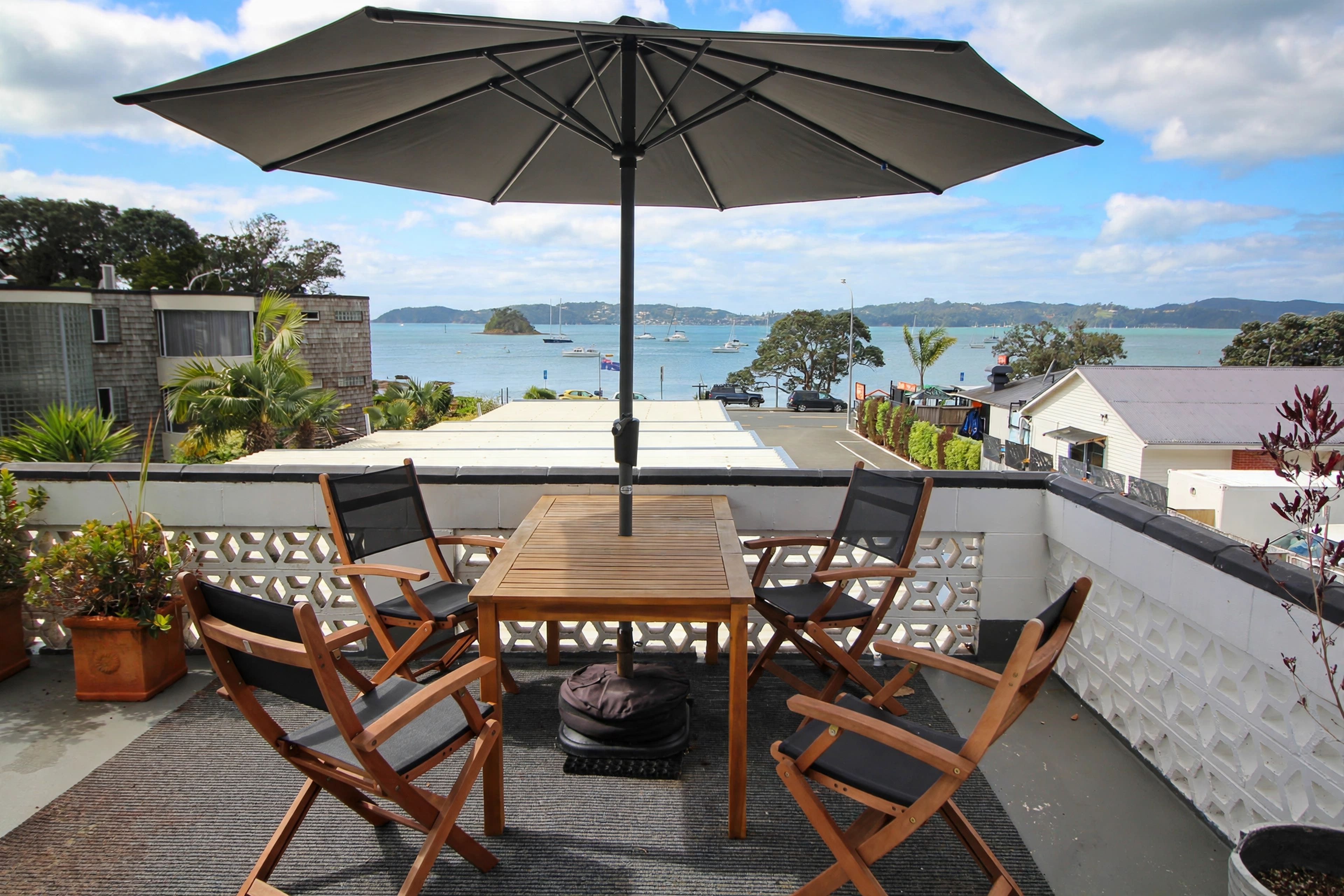 A wooden table and chairs with an umbrella, overlooking a harbor with boats and hills.