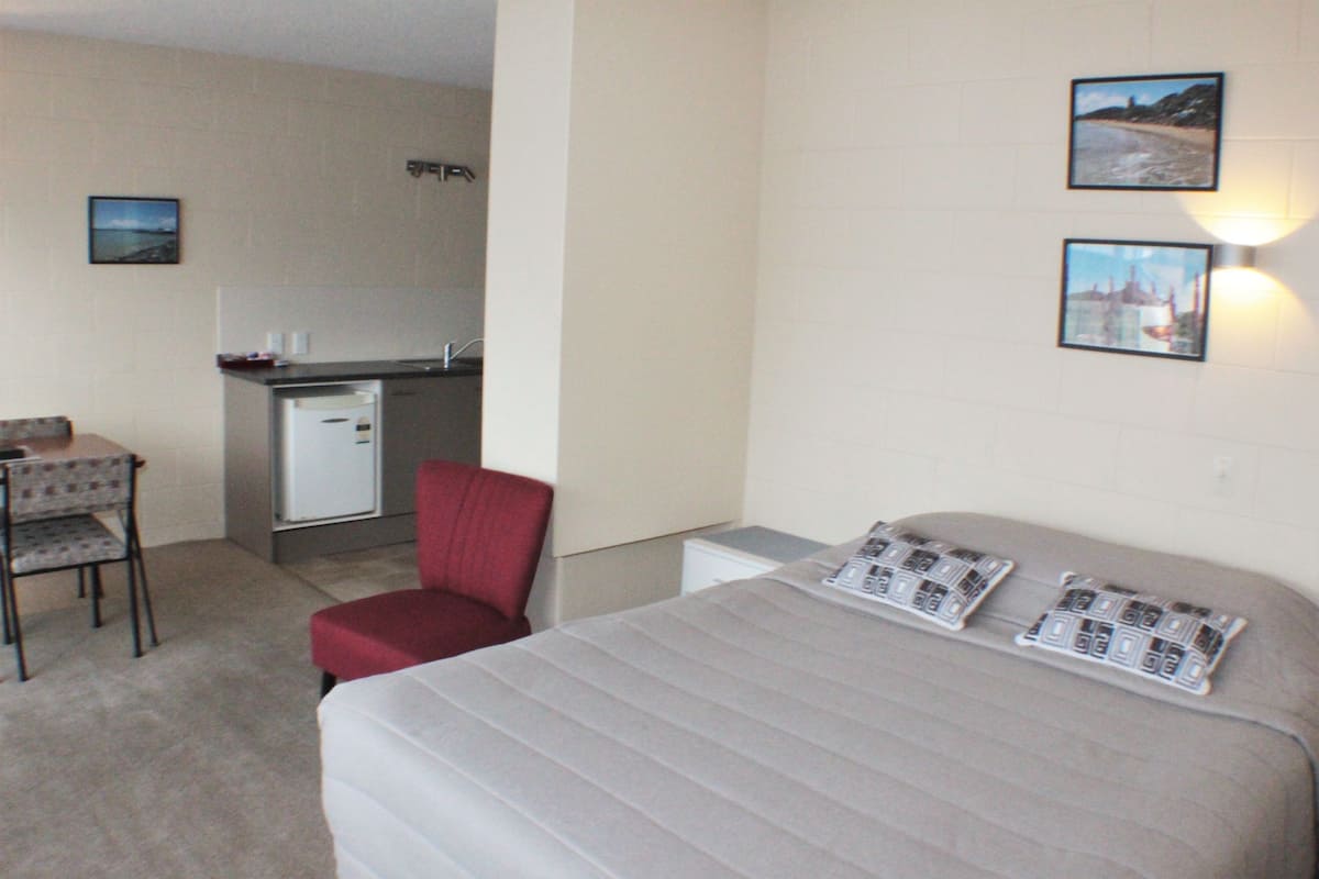 A hotel room with a bed, a chair, a small kitchenette, and two framed pictures on the wall.