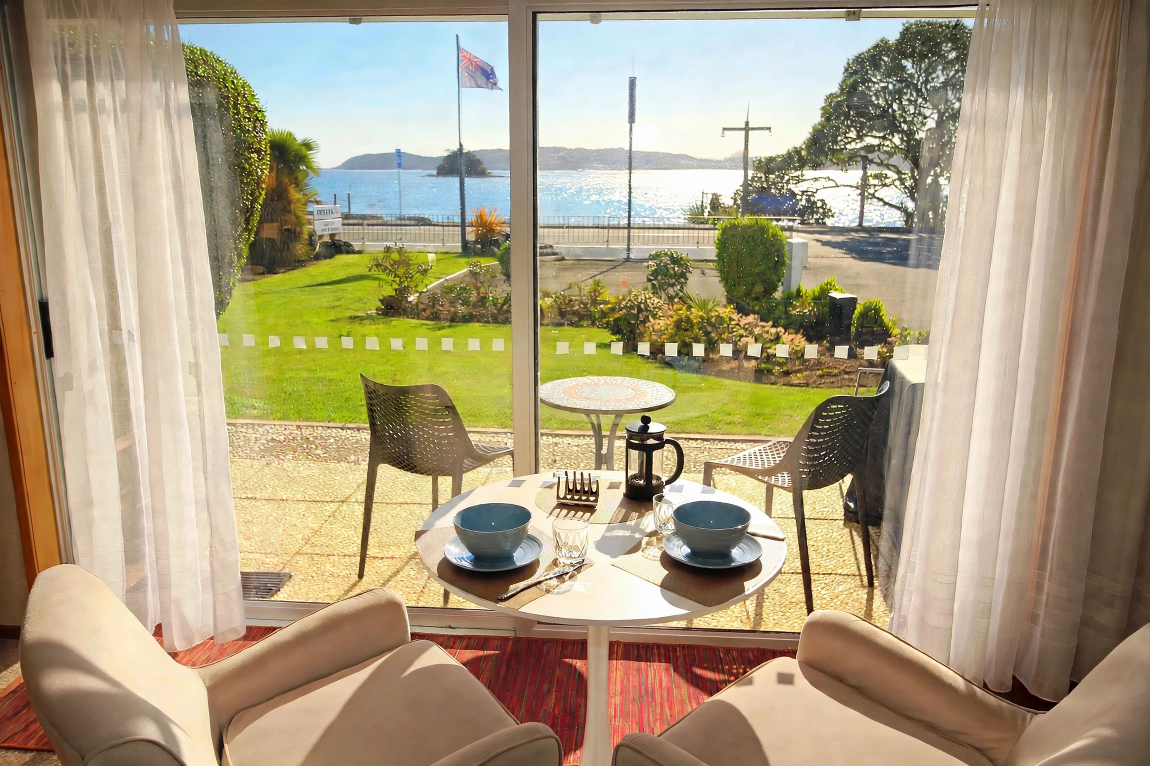 Habitación con vista a un jardín ajardinado y al mar. Cuenta con una mesa, sillas y una cafetera.