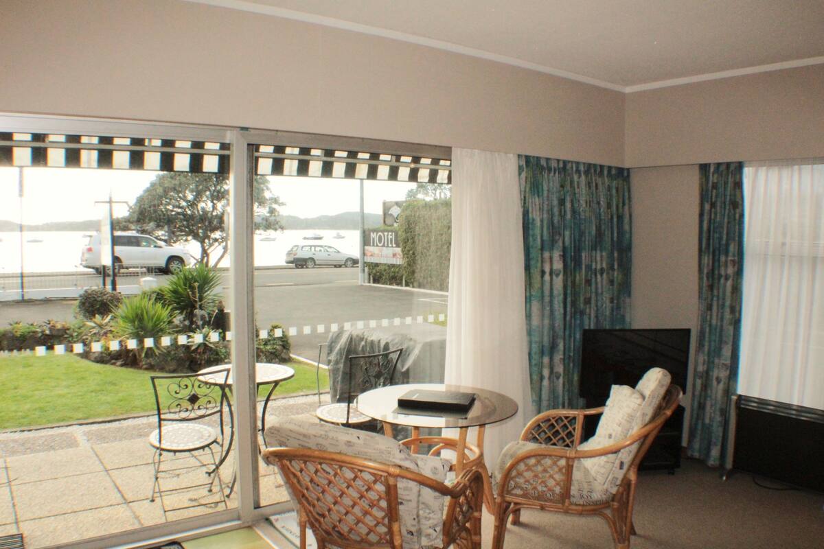A room with a balcony, wicker furniture, a television, and a view of the street and greenery outside.
