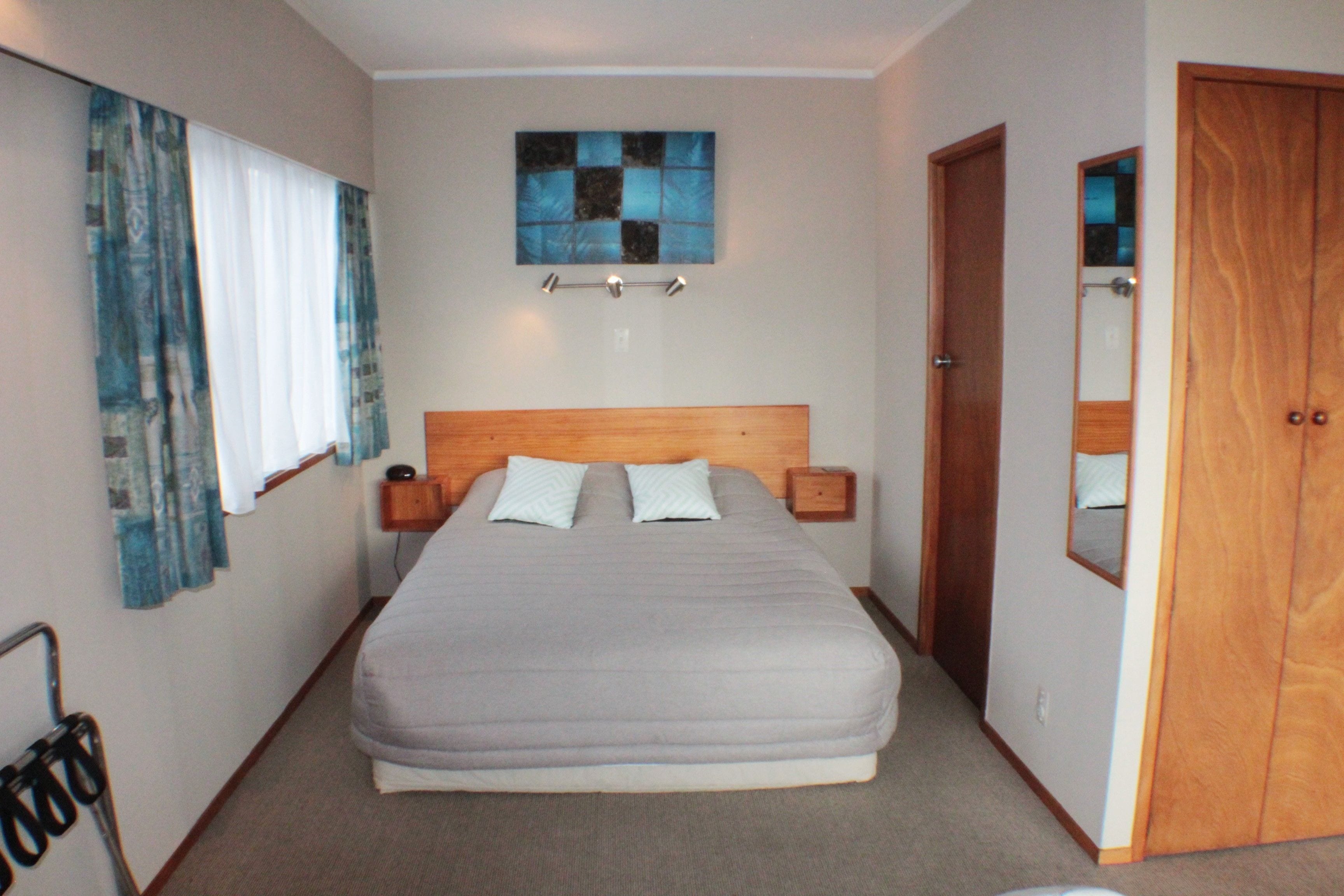 A neatly arranged bedroom with a large bed, bedside tables, a headboard, a wall-mounted light fixture, a window with curtains, and a wooden door.