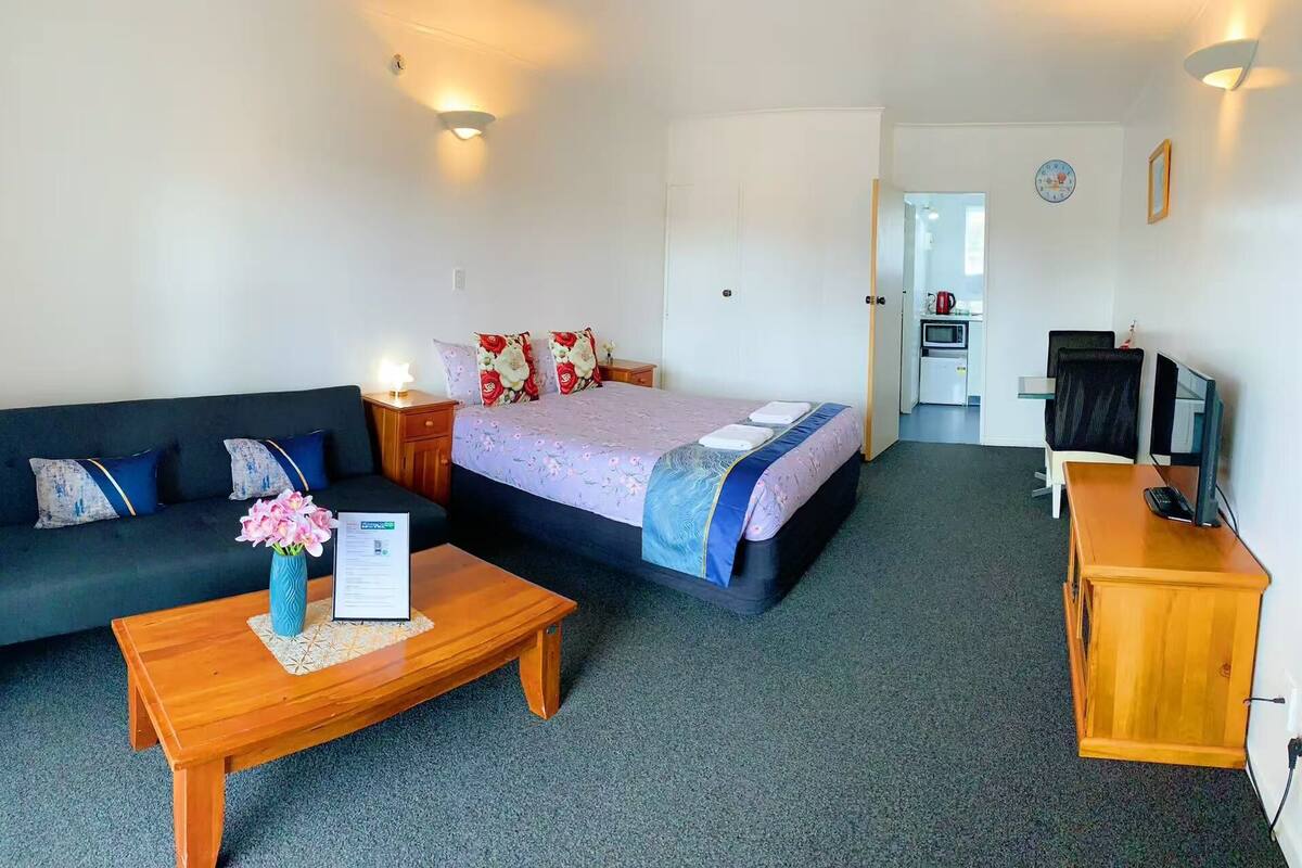 A hotel room with a bed, a sofa, a wooden table, a TV, and a kitchenette in the background.