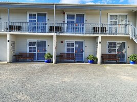 Een tweelaags motel met drie deuren, elk voorzien van een nummer. Er staan bankjes en potplanten voor de deuren.