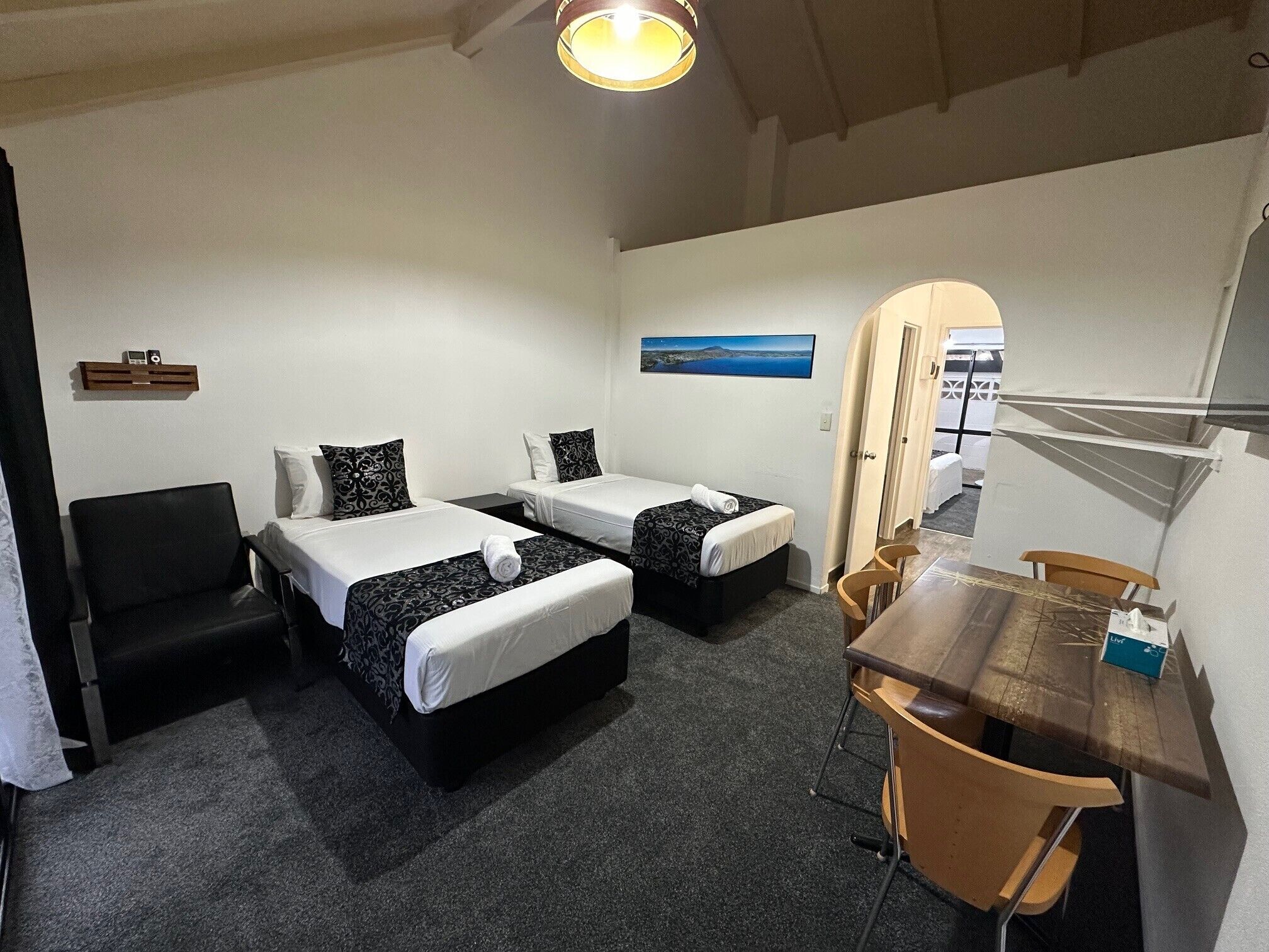 Ein Hotelzimmer mit zwei Betten, einem Schreibtisch, einem Stuhl, einem kleinen Tisch, einem Spiegel, einem Bild an der Wand und einer Deckenleuchte.