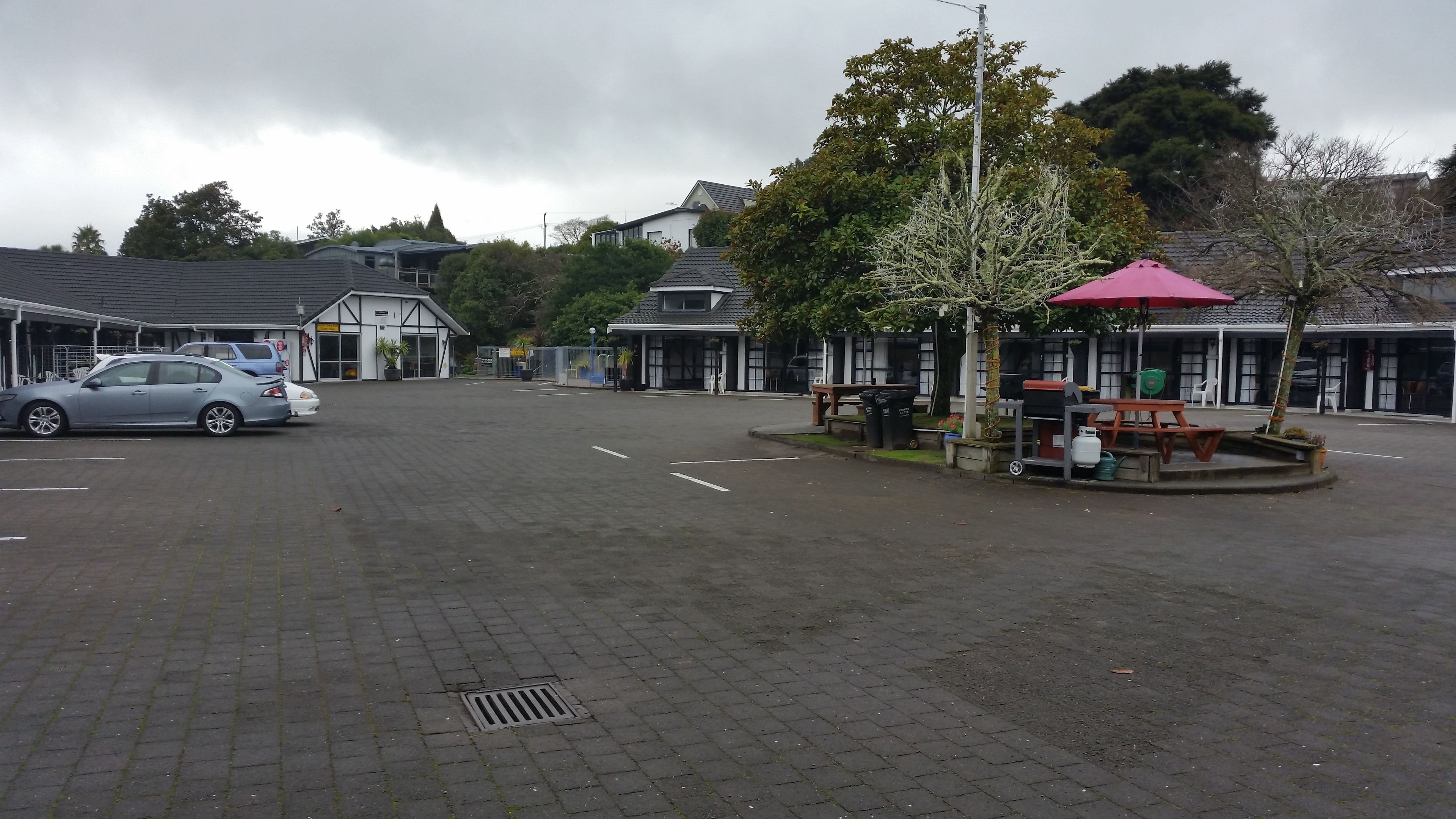 A parking lot with a car, a picnic area with a red umbrella, and buildings with large windows in the background.