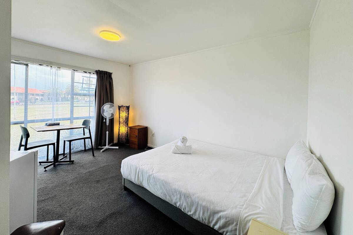 Une chambre d’hôtel équipée d’un lit, d’un bureau, d’une chaise, d’un ventilateur sur pied et d’une fenêtre donnant sur l’extérieur.