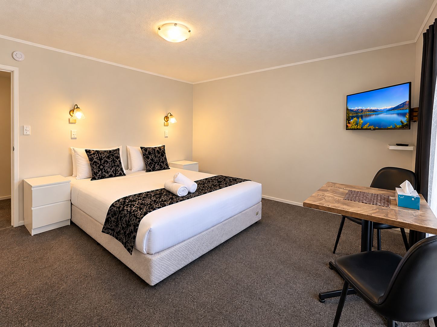 A hotel room with a bed, bedside tables, a desk with a chair, a television, and a view of a lake.