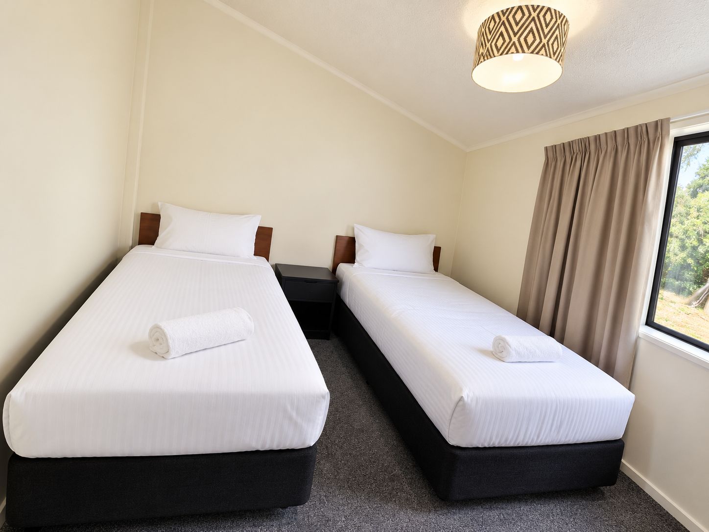 A hotel room with two beds, a window with curtains, and a patterned ceiling light.